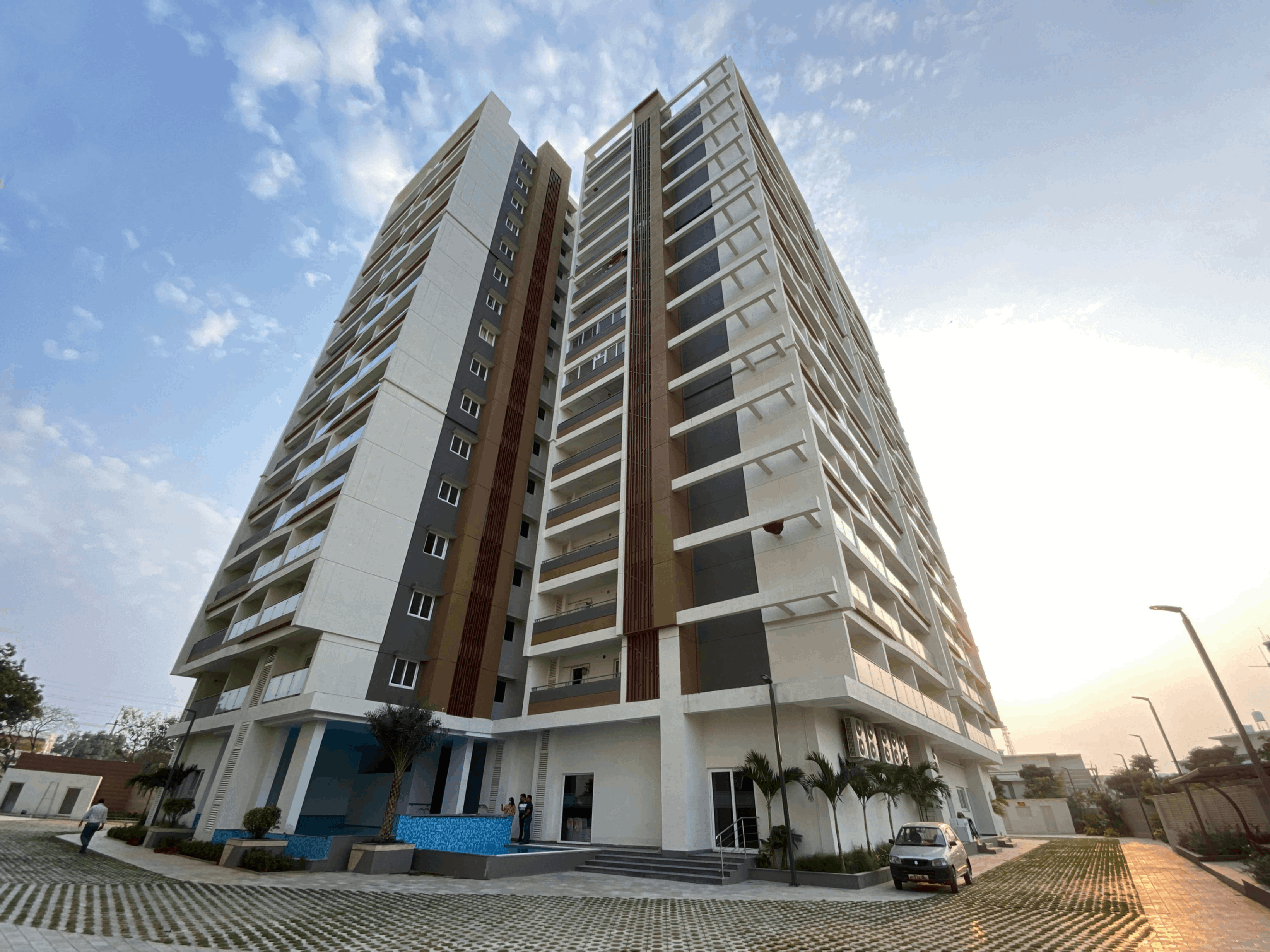 Modern multi-story residential building with balconies, surrounded by paved walkways and landscaped greenery under a partly cloudy sky at sunset.
