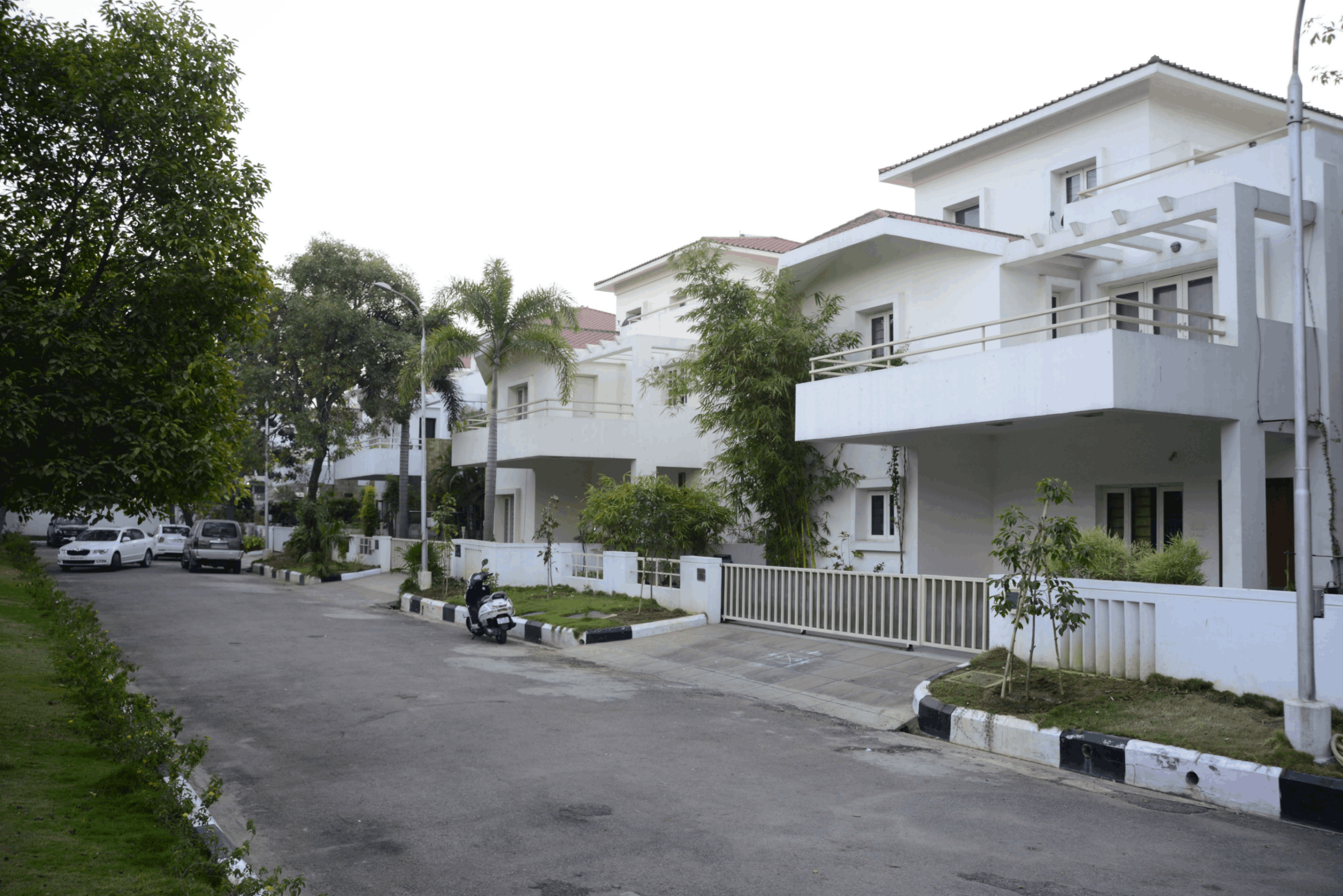 Modern white two-story houses with balconies along a quiet street lined with trees and parked cars.