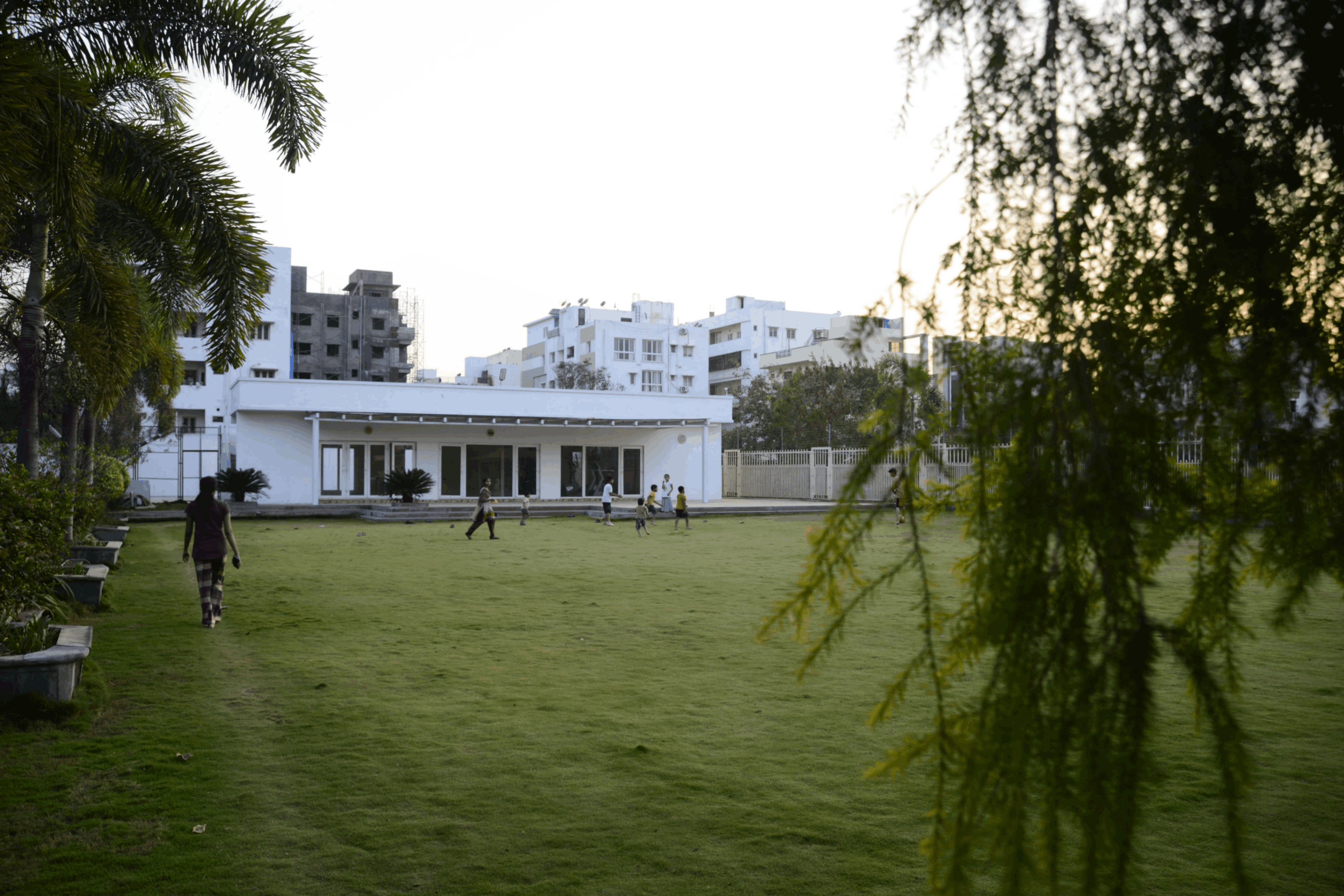 People walking and children playing on a large grassy lawn in front of a white building with urban buildings in the background.