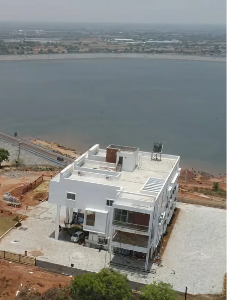 Aerial view of a modern, white, multi-story building near a large body of water with surrounding dry land and a distant road.