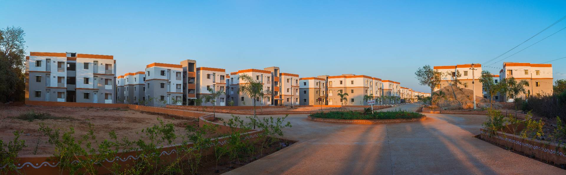 Row of white and orange modern three-story apartment buildings with a paved road and landscaped plants in front under a clear blue sky.
