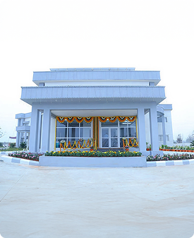 Modern white building decorated with yellow and orange flower garlands, surrounded by flower beds and a paved area.