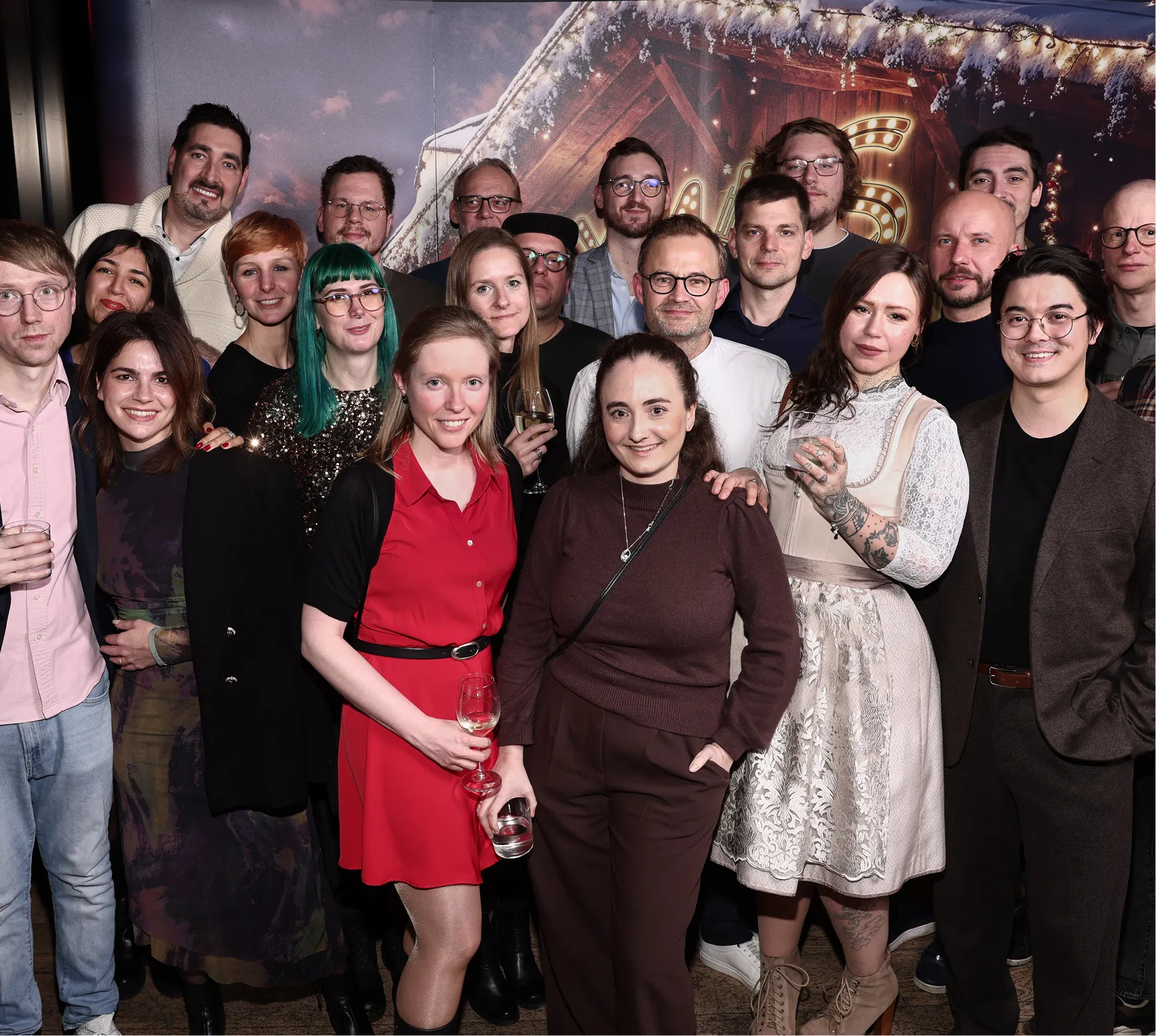 Gruppenfoto von 22 lächelnden Erwachsenen bei einer Feier, mit Getränken in der Hand und einem festlich dekorierten Hintergrund.