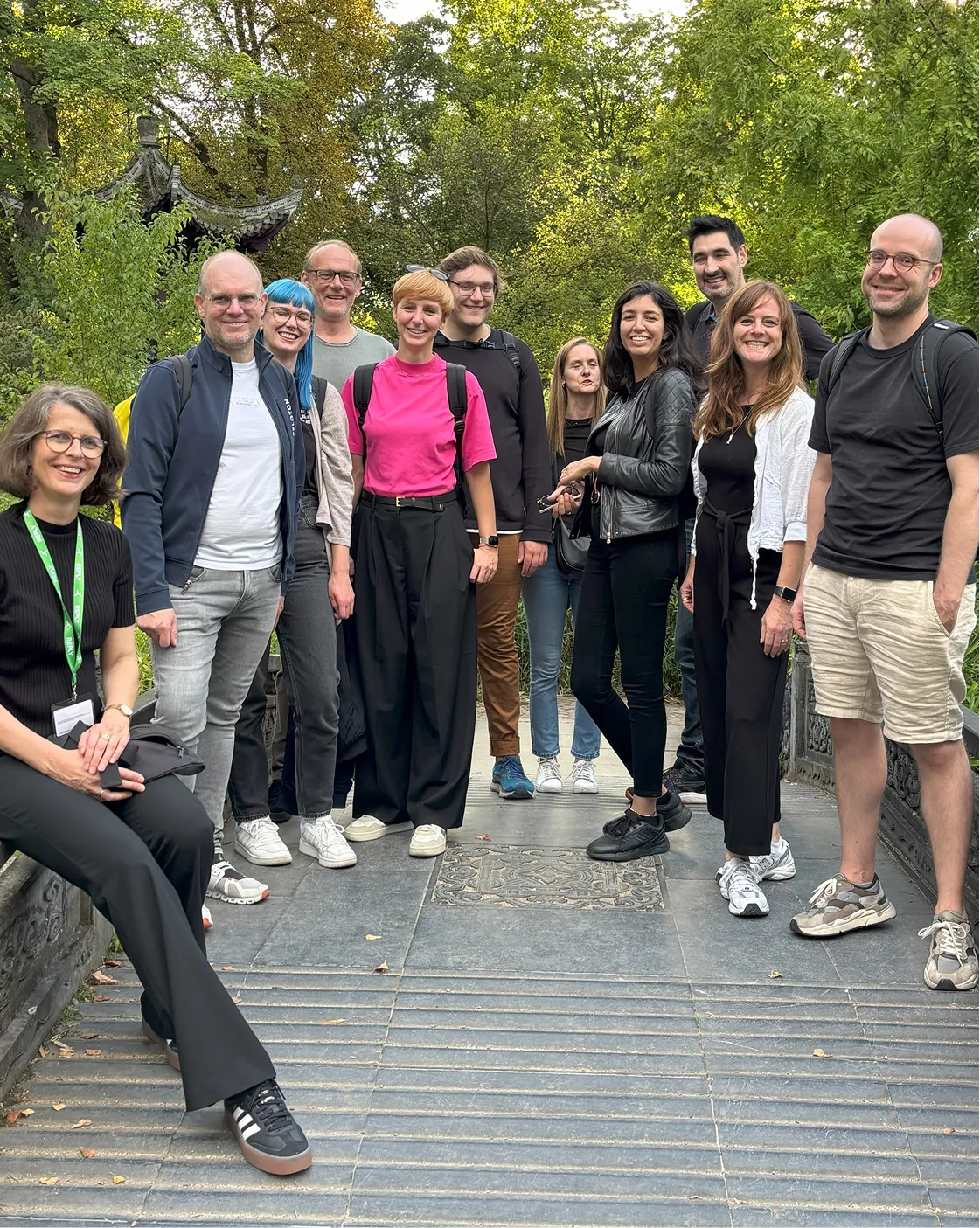 Eine Gruppe von zehn Menschen steht auf einer Brücke in einem grünen Park, alle lächeln in die Kamera.