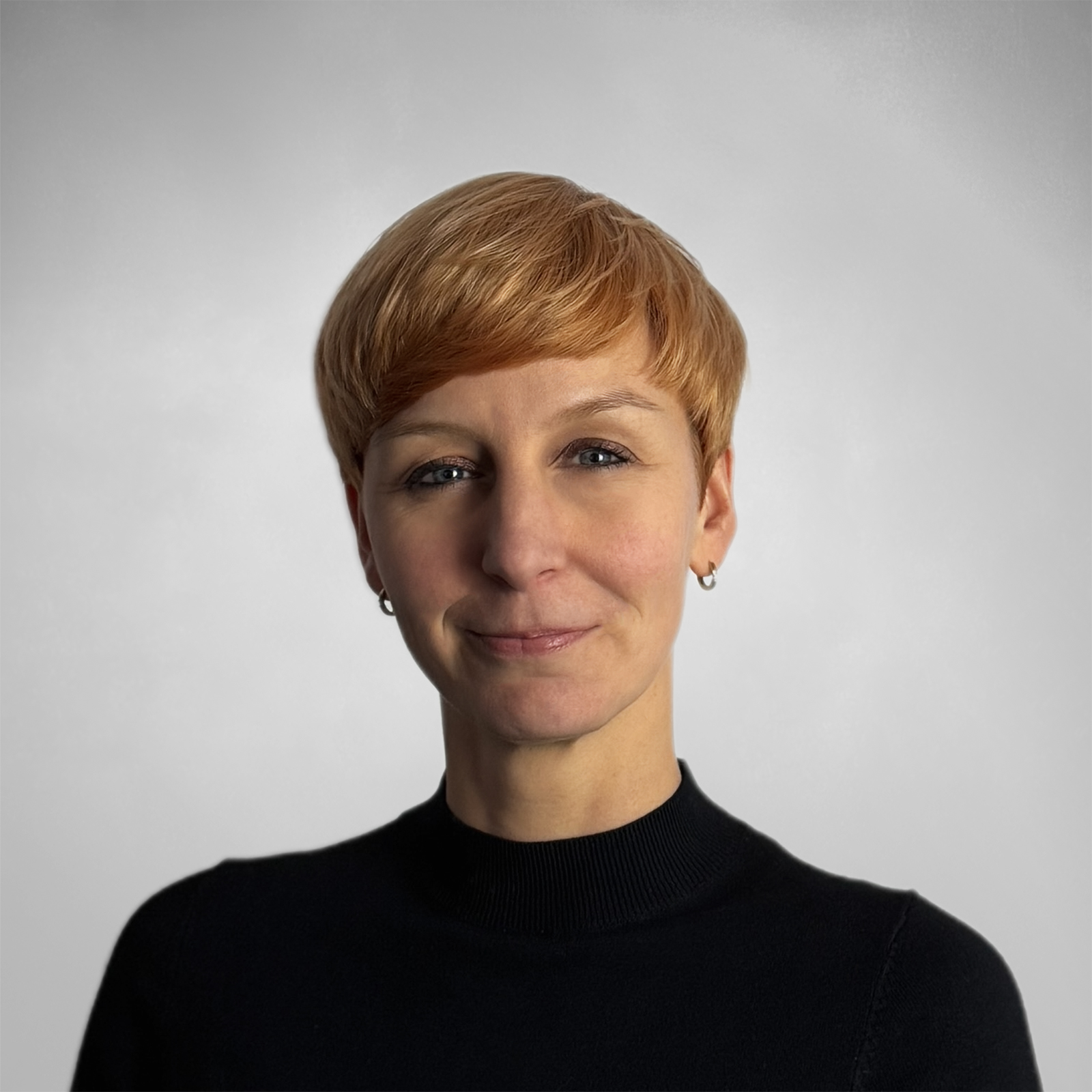 Porträt einer Frau mit kurzem roten Haar, die ein schwarzes Oberteil trägt und leicht lächelt vor einem hellgrauen Hintergrund.