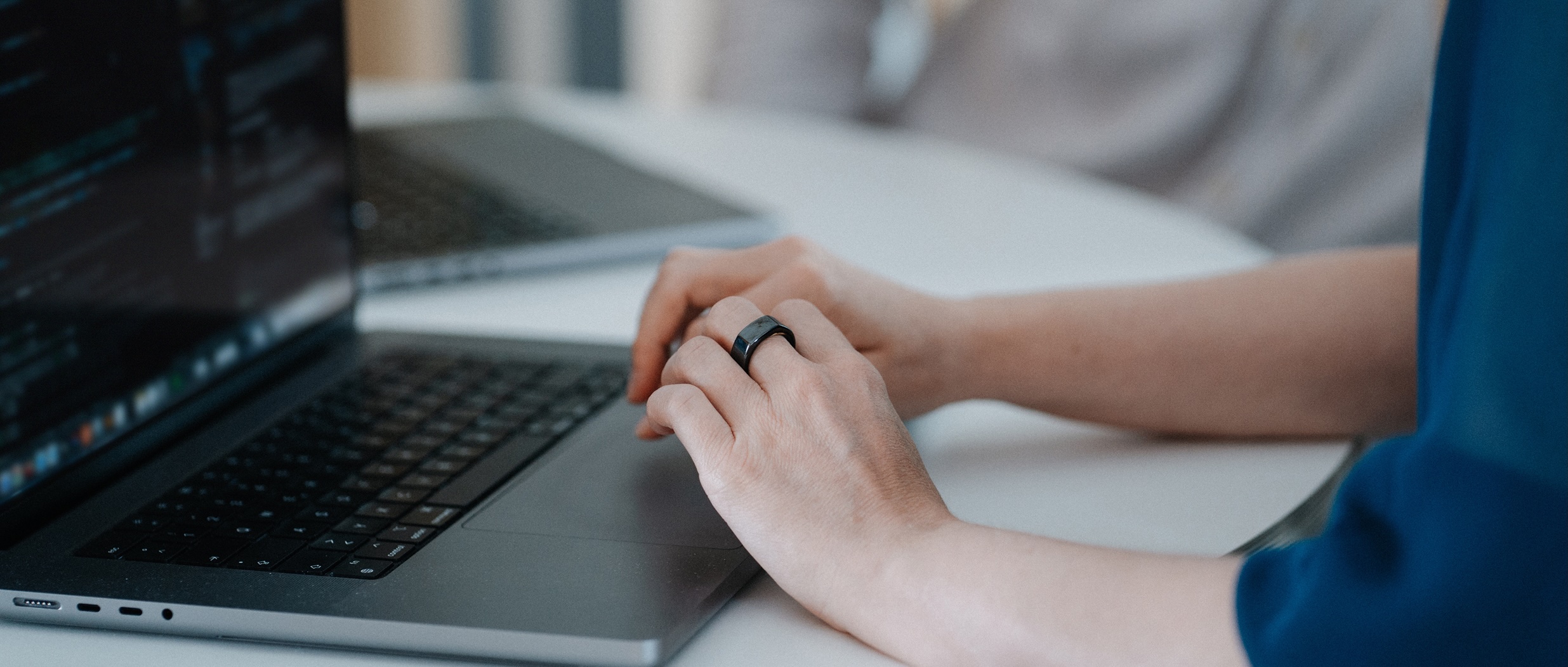 Hände mit einem schwarzen Ring tippen auf der Tastatur eines Laptops, im Hintergrund ein weiterer Laptop auf einem weißen Tisch.
