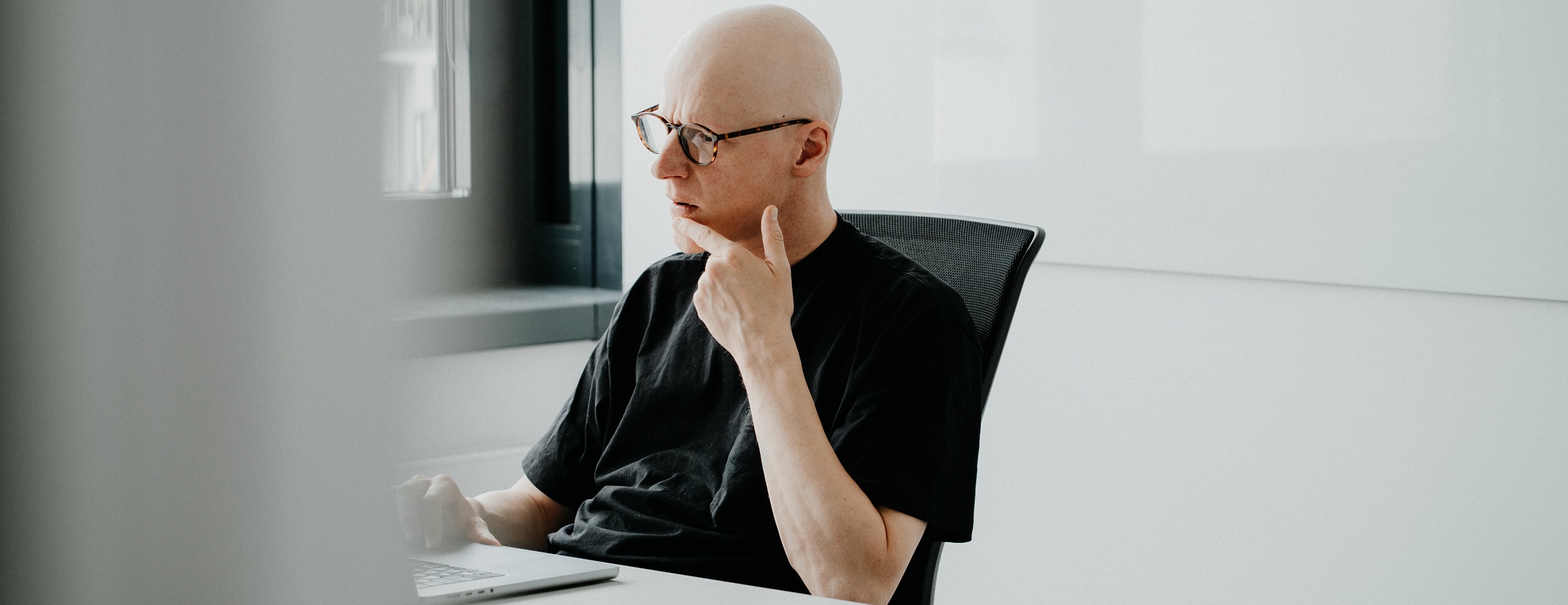 Ein nachdenklicher Mann mit Brille und schwarzem T-Shirt sitzt an einem Schreibtisch vor einem Laptop in einem hellen Raum.