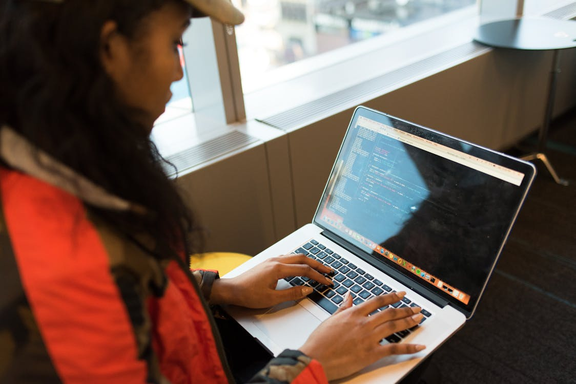 A person in a red jacket and cap typing on a laptop, with lines of code visible on the screen in front of a window