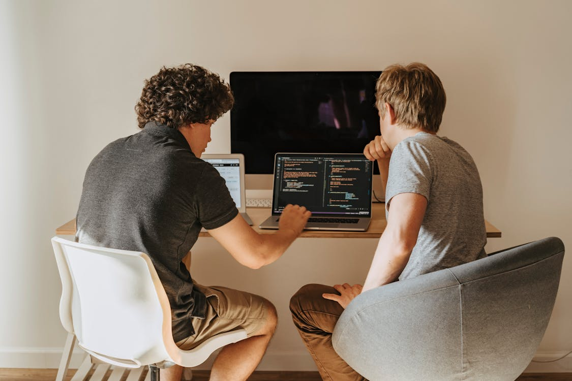 Two people sitting at a desk collaborating on coding projects, both looking at laptops with code on the screens