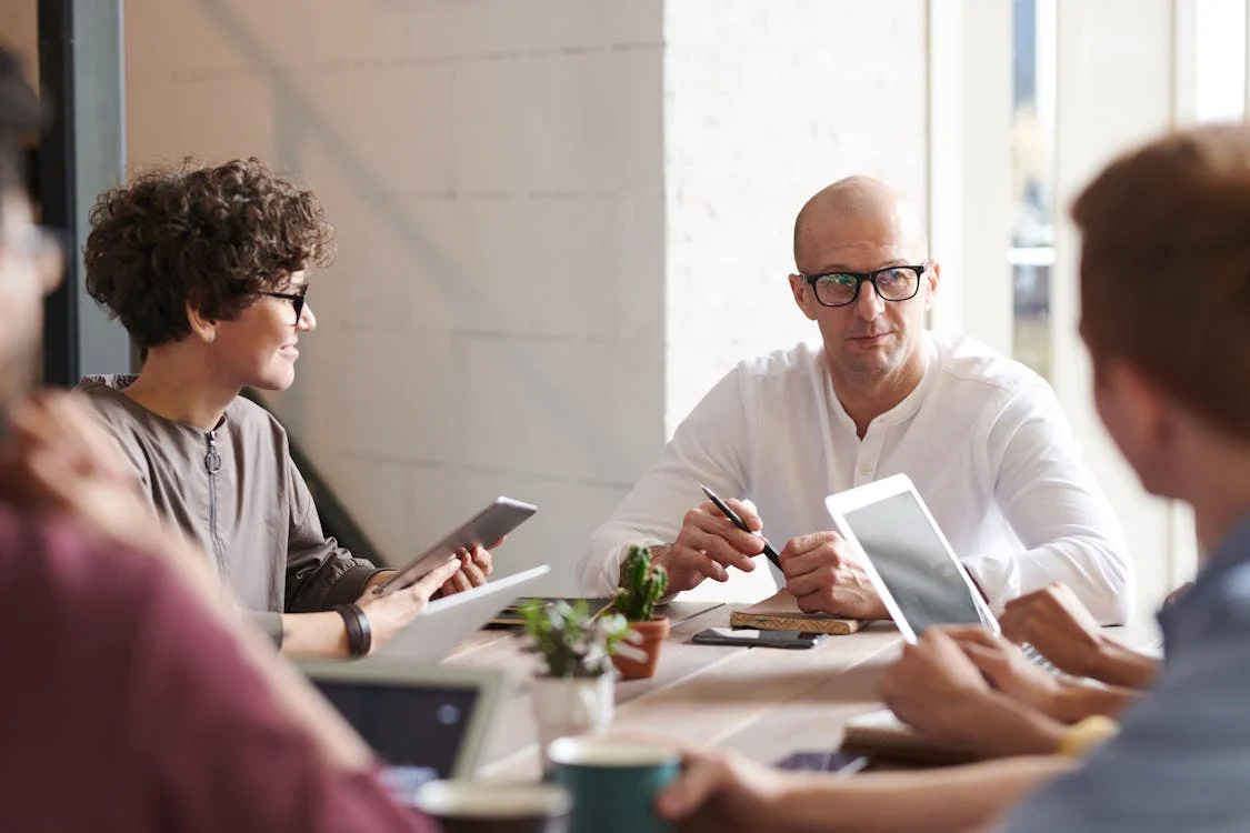 A group of professionals in a meeting, discussing around a table with laptops and tablets