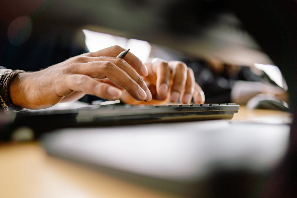 Close-up of a person’s hands typing on a keyboard with a pen in hand