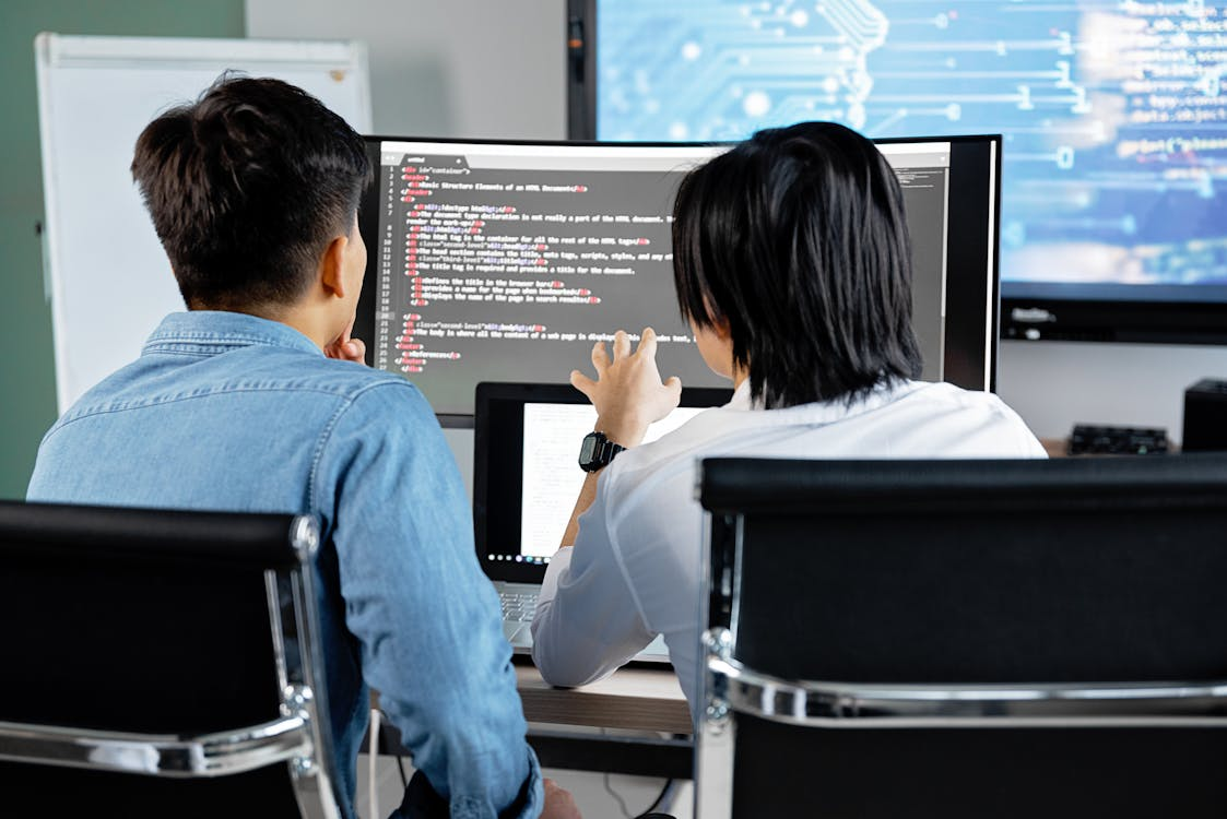 Two developers collaborating while reviewing code on a large dual-screen monitor, discussing AI threat hunting findings