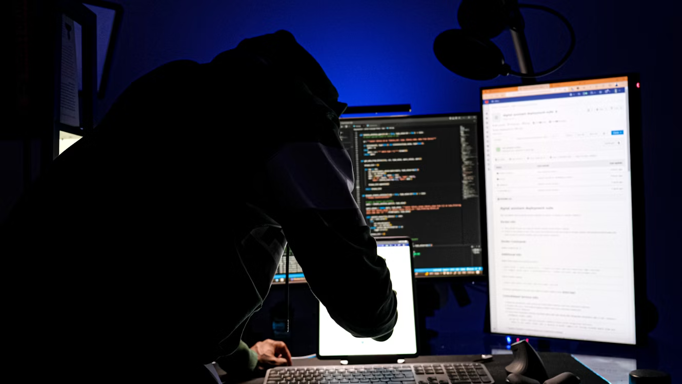 Person in a dark room working at a multi-monitor computer setup, suggesting a cybersecurity threat or malicious activity