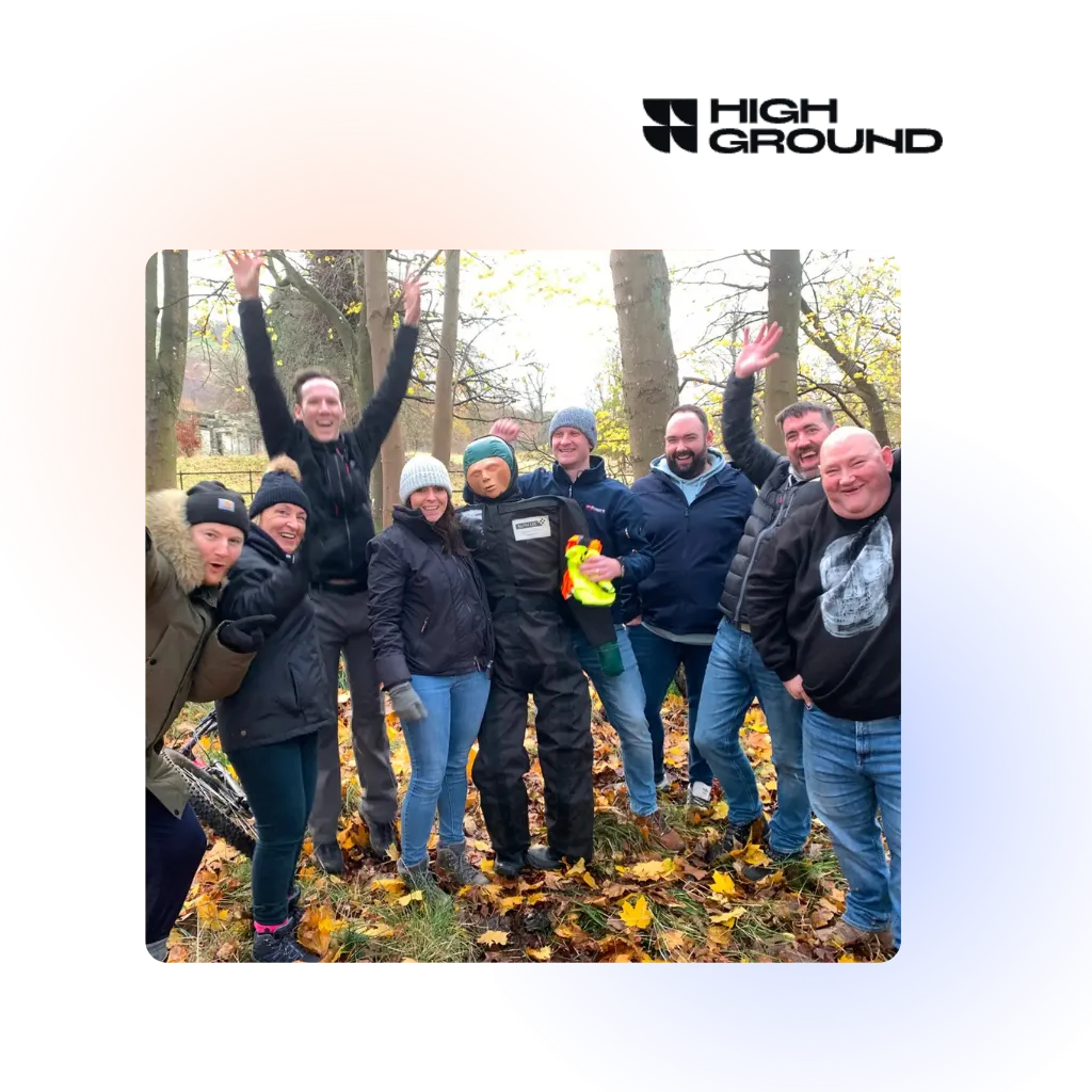 Group of eight people, one jumping with arms raised, standing outdoors among fall foliage, with one person holding a training dummy.