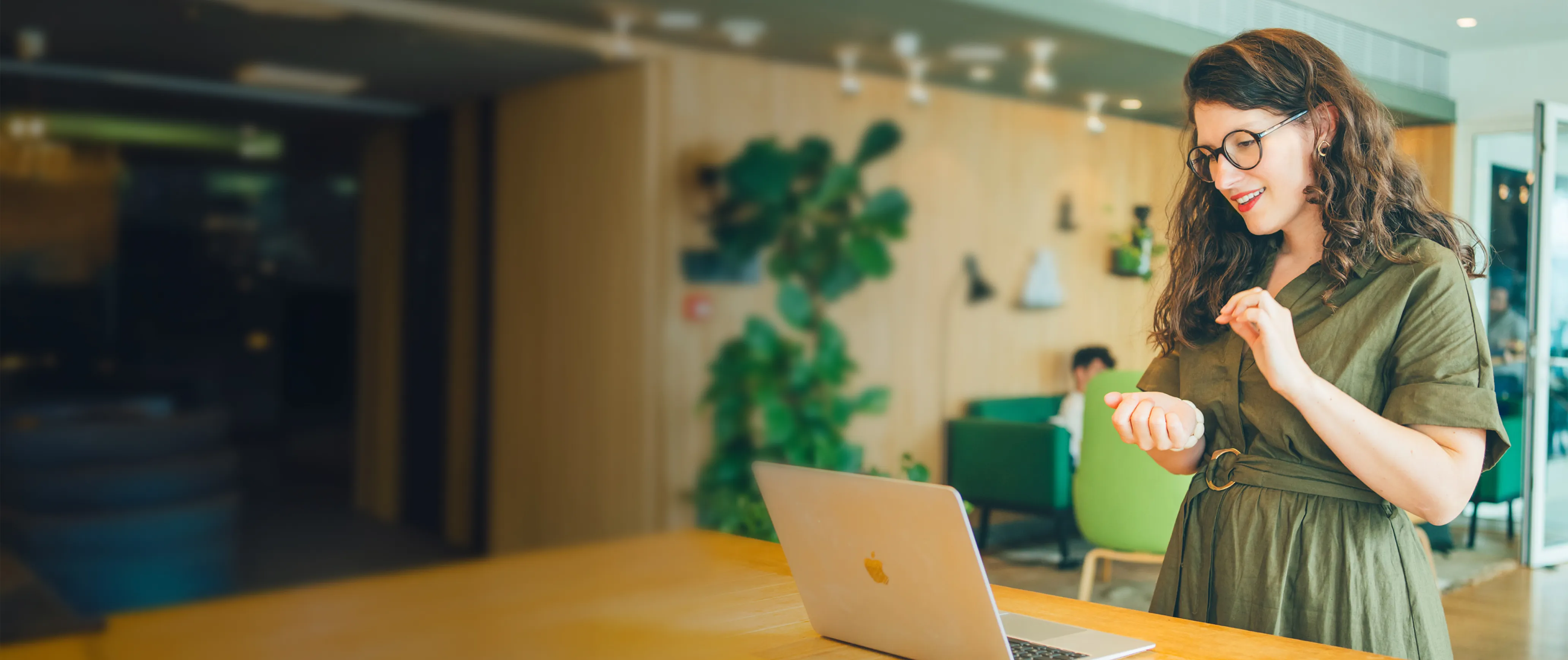 Une femme souriante en robe verte, portant des lunettes, se tient debout devant un ordinateur portable posé sur une table en bois dans un espace de travail moderne et lumineux.