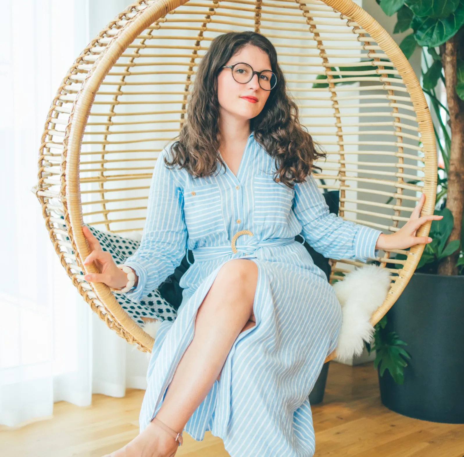 Une femme assise dans un fauteuil suspendu en osier, portant une robe bleue à rayures, des lunettes rondes, et faisant le signe de la paix avec ses mains.