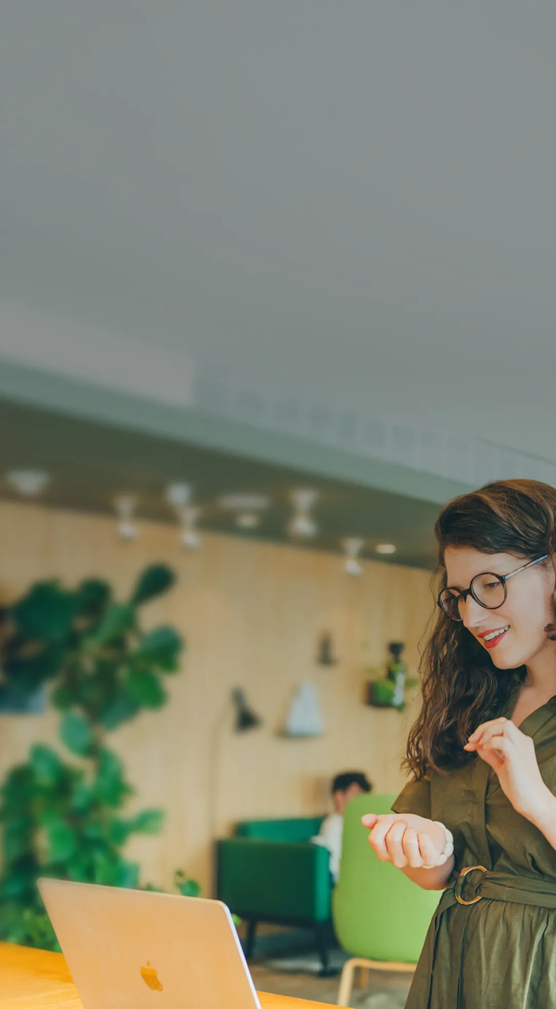 Une femme souriante portant des lunettes rondes et une robe verte, debout devant un ordinateur portable posé sur une table en bois dans un espace de travail moderne et lumineux.