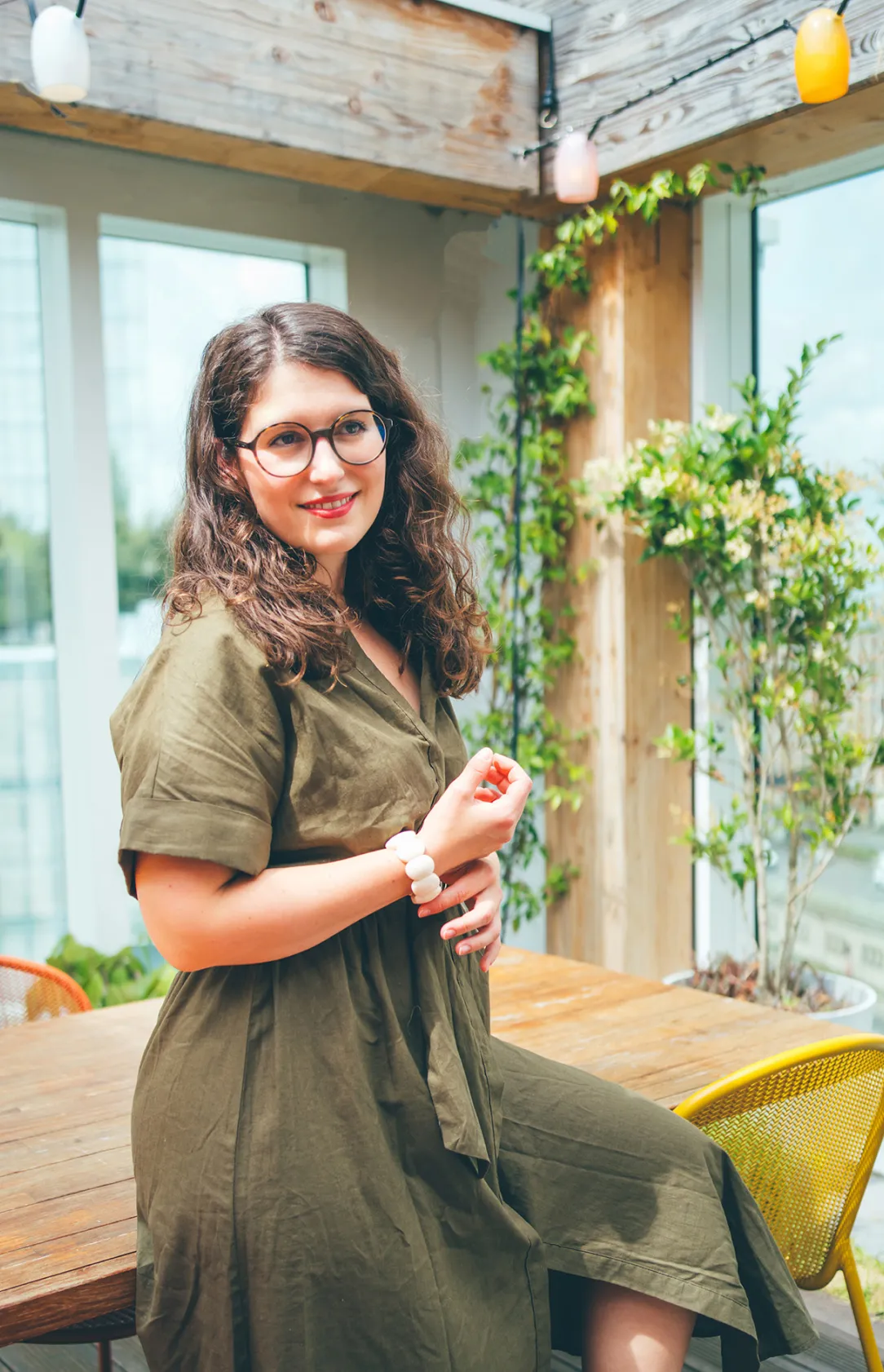 Une femme souriante portant des lunettes rondes et une robe verte, assise sur une table en bois dans un espace extérieur lumineux avec des plantes en arrière-plan.