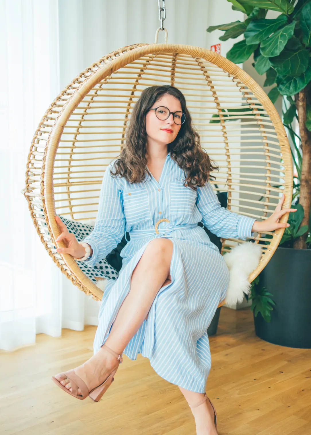 Une femme assise dans un fauteuil suspendu en osier, portant une robe bleue à rayures, des lunettes rondes, et faisant le signe de la paix avec ses mains.