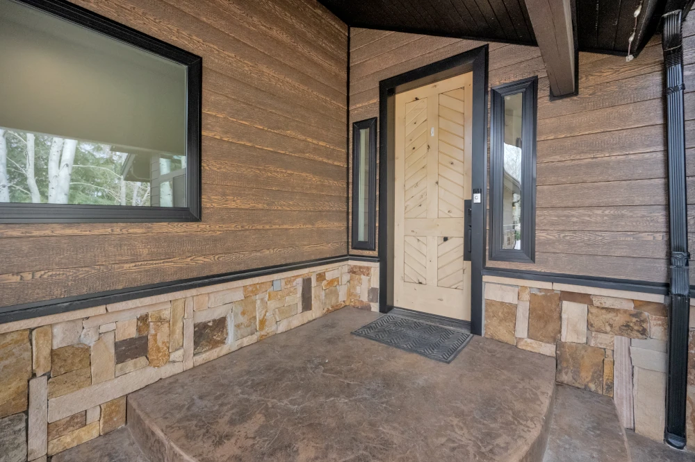 Covered entryway with a natural wood chevron front door, black-framed sidelights, and stacked stone wainscot
