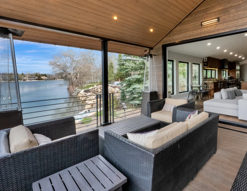 Covered lakeside deck with wood plank ceiling, recessed lighting, wicker patio seating, and folding glass doors opening to the interior