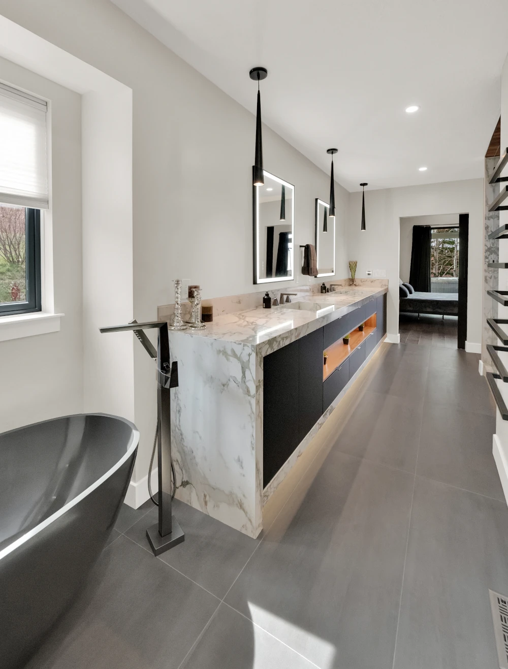 Master bathroom with marble waterfall double vanity, backlit mirrors, black pendant lights, freestanding black soaking tub, and large-format gray tile flooring