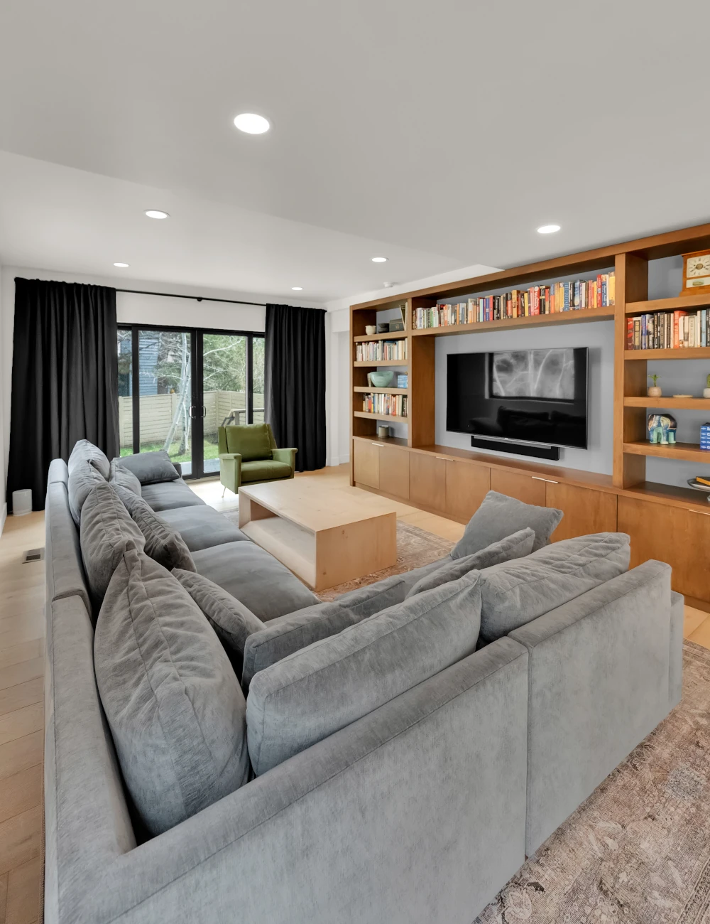 Remodeled living room with a gray sectional sofa, built-in wood bookshelves and media cabinetry, wall-mounted TV, and sliding glass patio doors