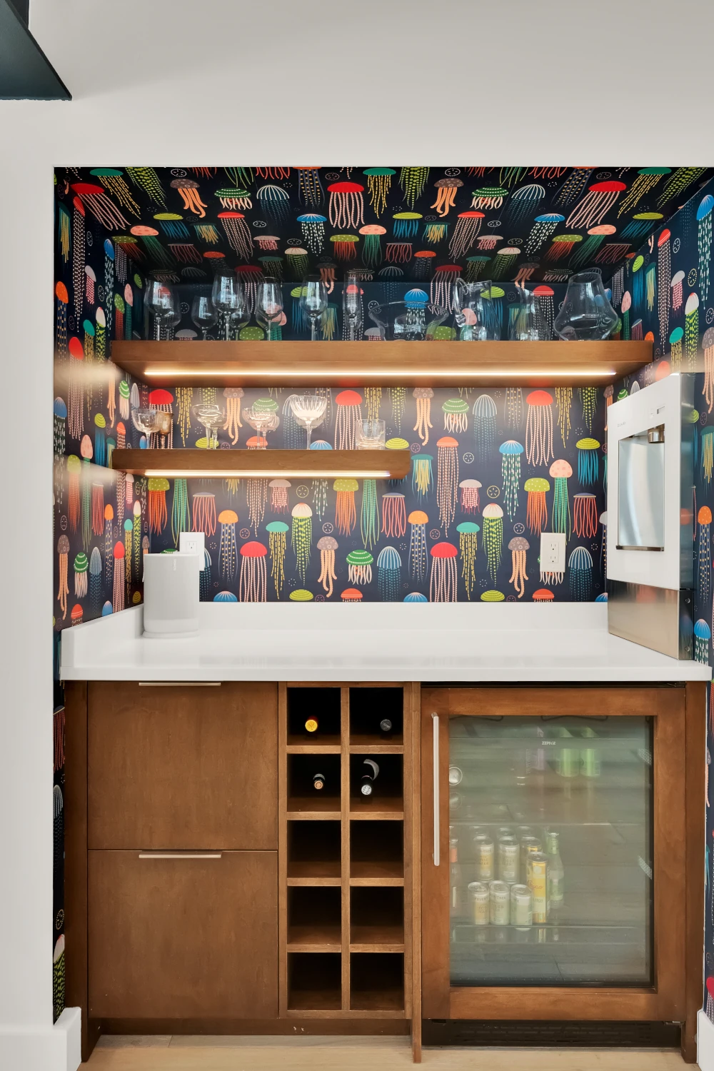 Built-in wet bar with wood cabinetry, wine storage cubbies, glass-front beverage fridge, lit floating shelves, and colorful jellyfish print wallpaper