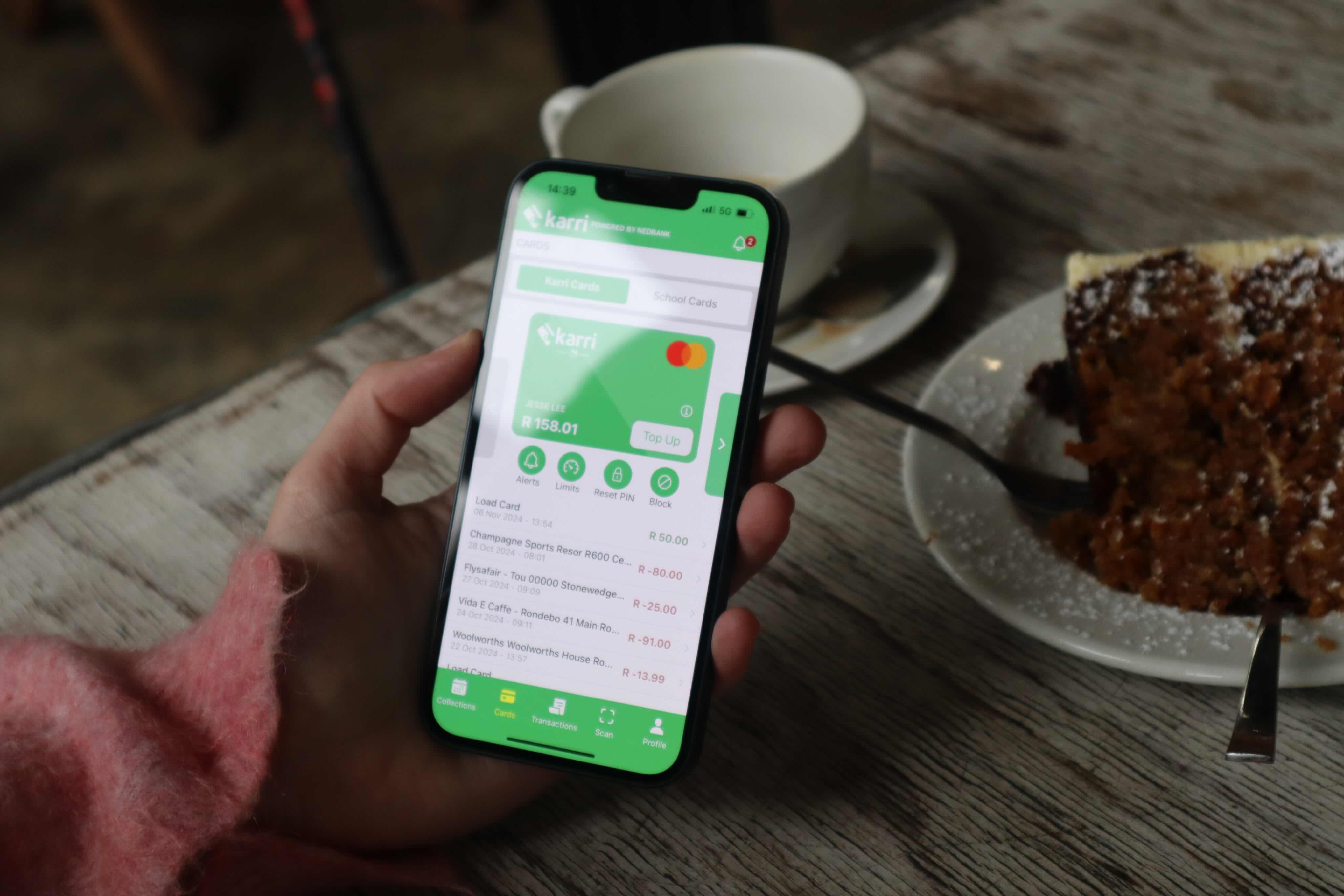 Hand holding phone displaying Karri app with balance and card options, next to coffee and cake on table.