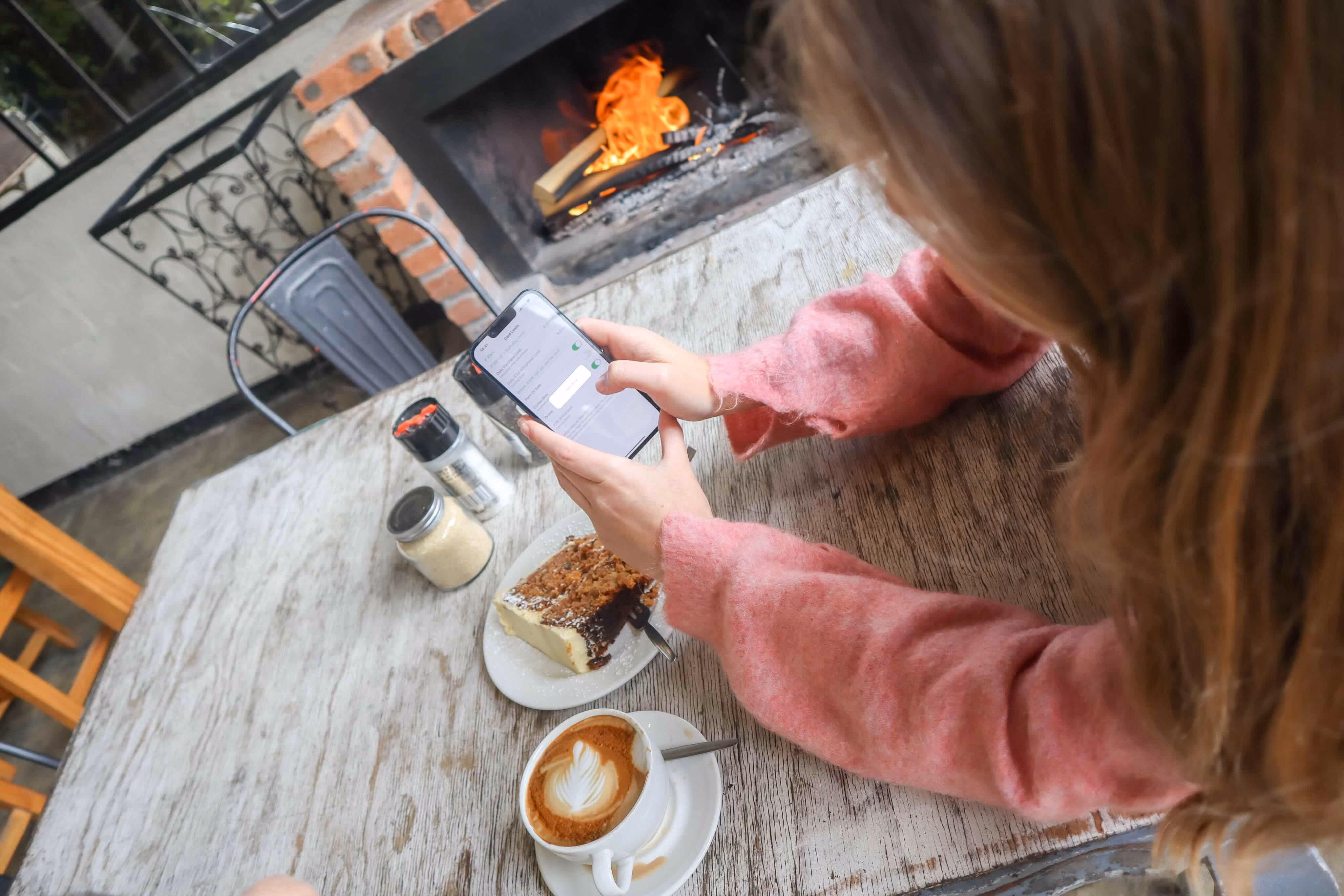 Person in pink sweater using a smartphone at a table with coffee, cake, and a fireplace nearby.