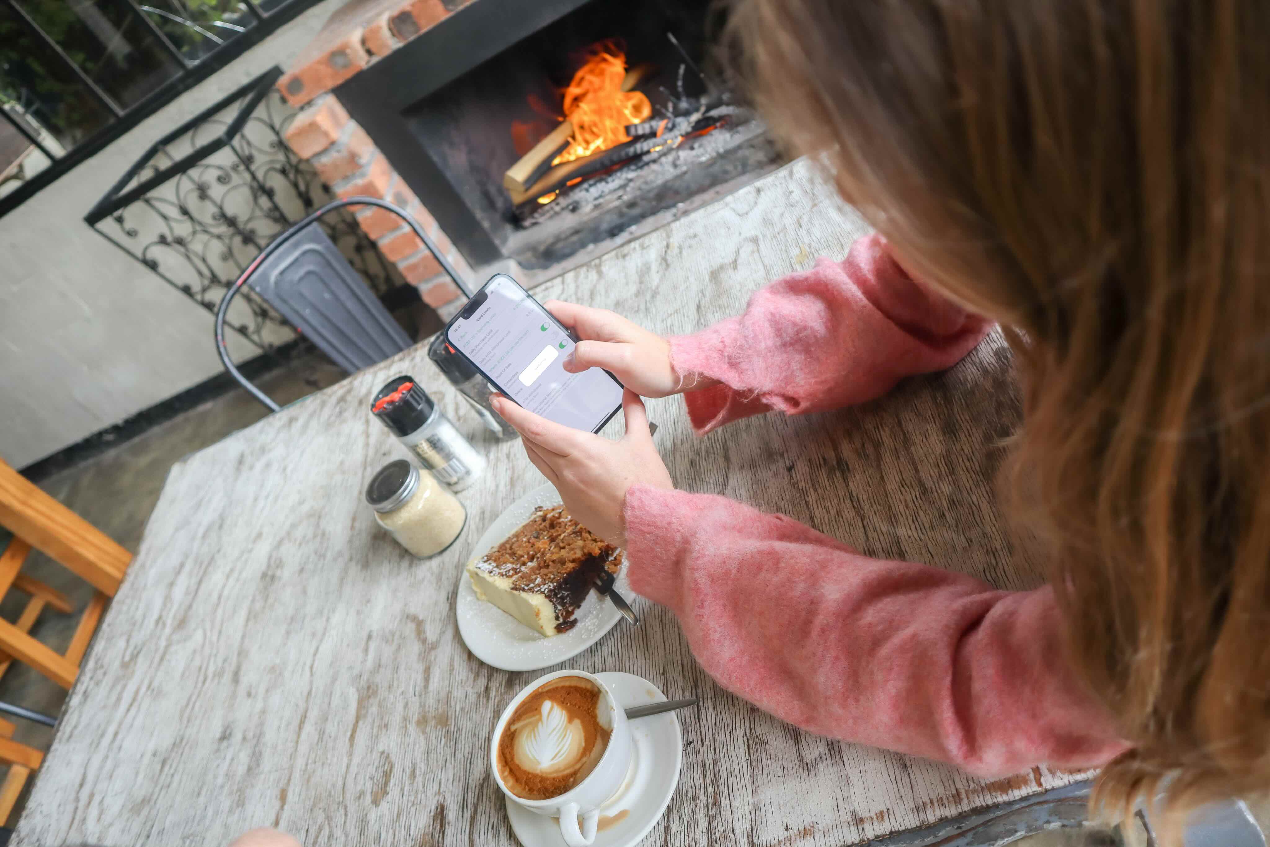 Person in pink sweater using a smartphone at a table with coffee, cake, and a fireplace nearby.
