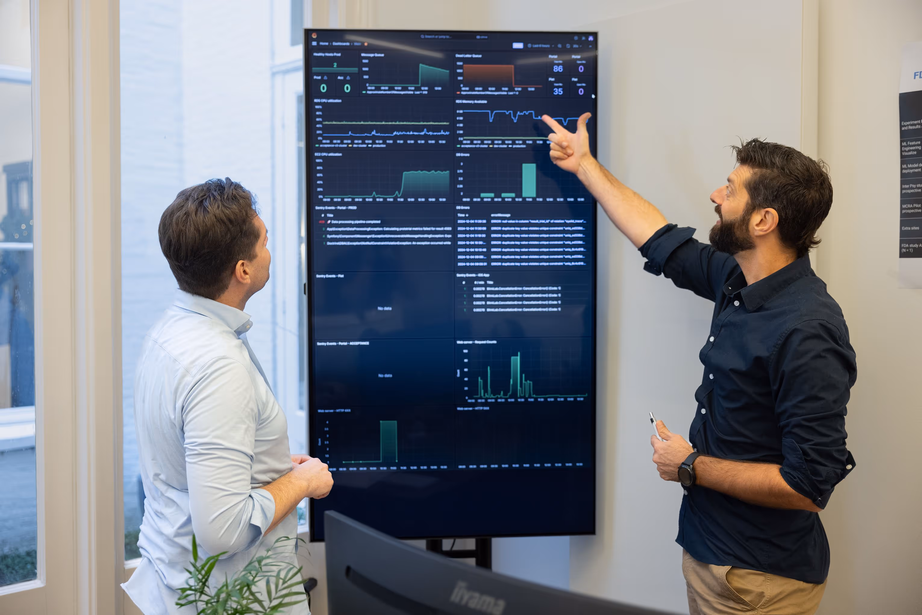 Two men in an office discussing data displayed on a large vertical screen with graphs and charts.