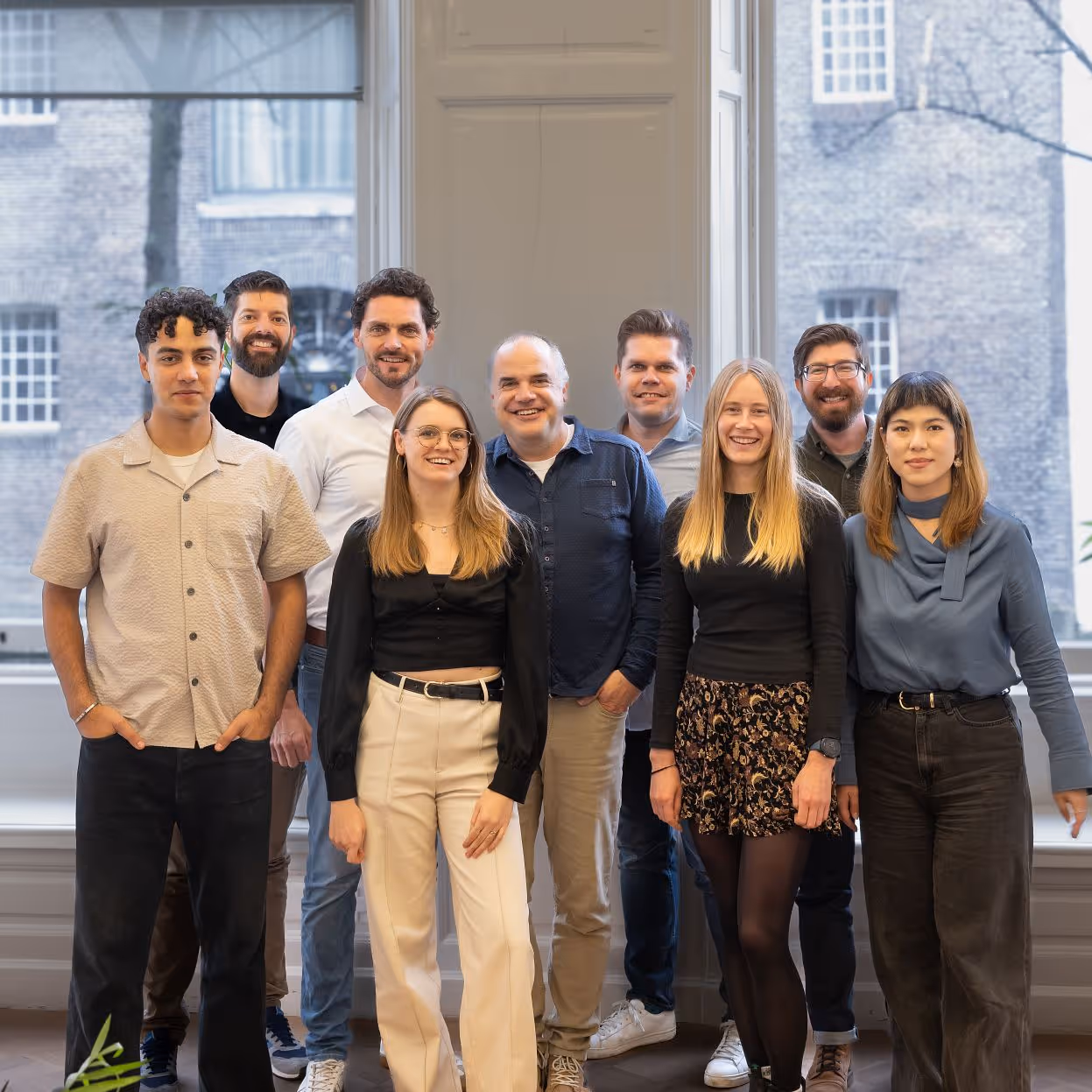 Group portrait of nine diverse adults standing indoors in front of large windows with brick building visible outside.