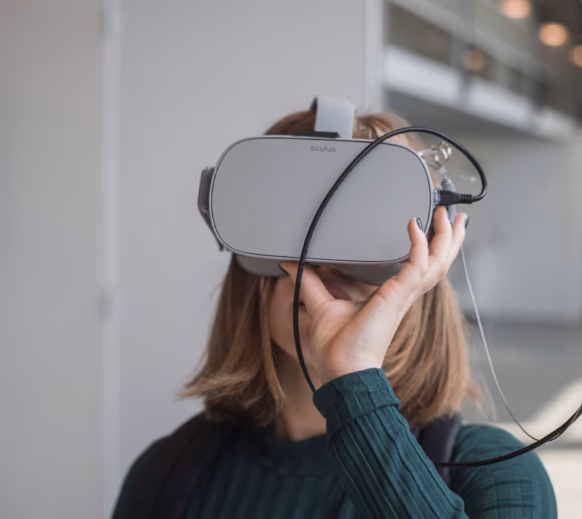 Person with shoulder-length hair wearing an Oculus virtual reality headset and holding a cable connected to it.