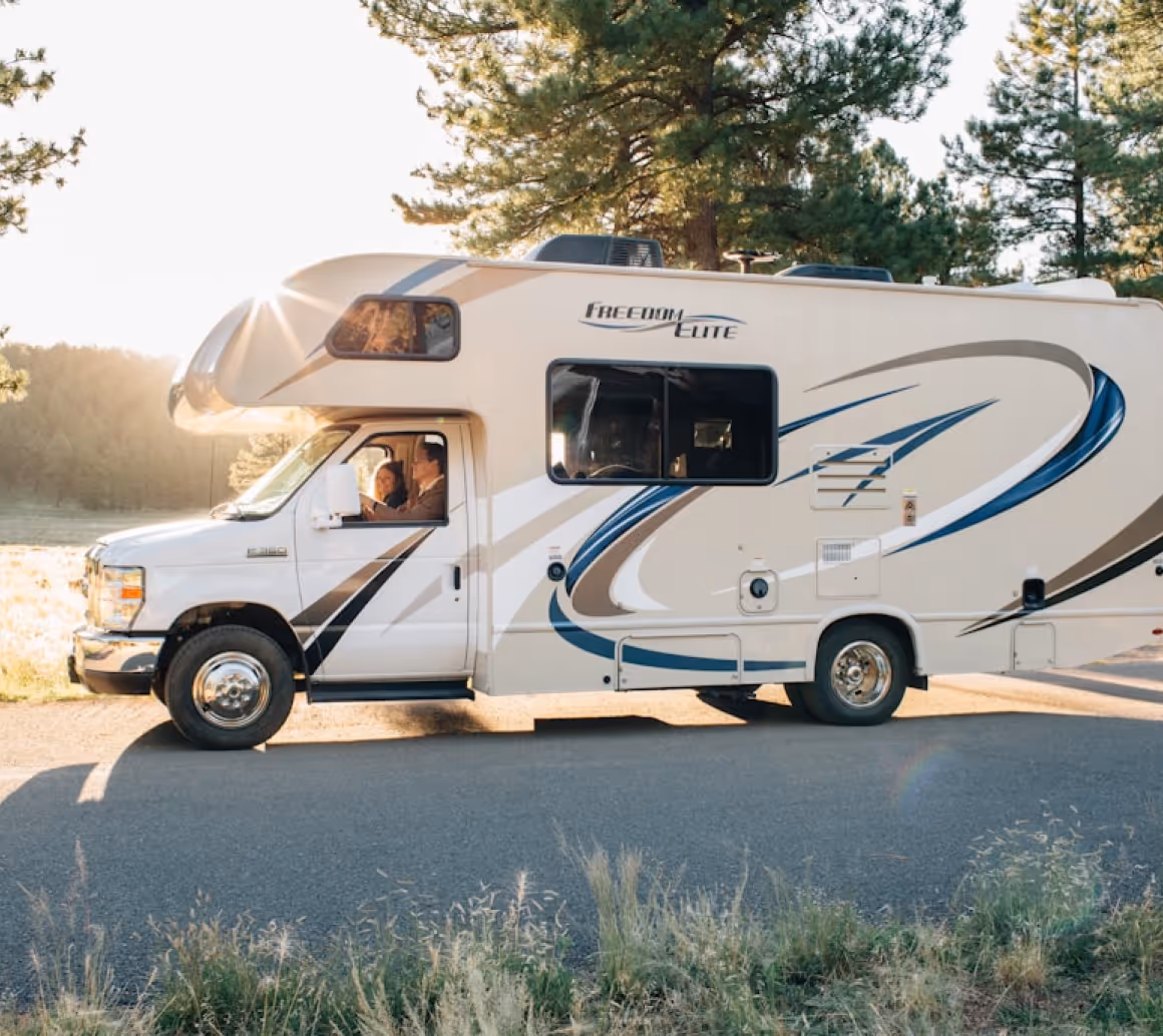White Freedom Elite motorhome parked on a road with two people visible inside and pine trees in the background.