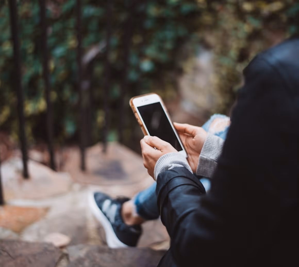 Person wearing a black jacket and ripped jeans sitting outdoors, holding a smartphone with a blank screen.