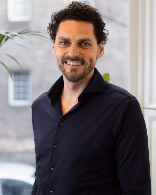 Smiling man with dark curly hair and beard wearing a black button-up shirt standing indoors near a window.