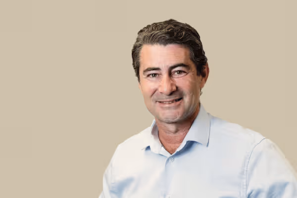 Middle-aged man with dark hair, wearing a light blue button-up shirt, smiling against a beige background.