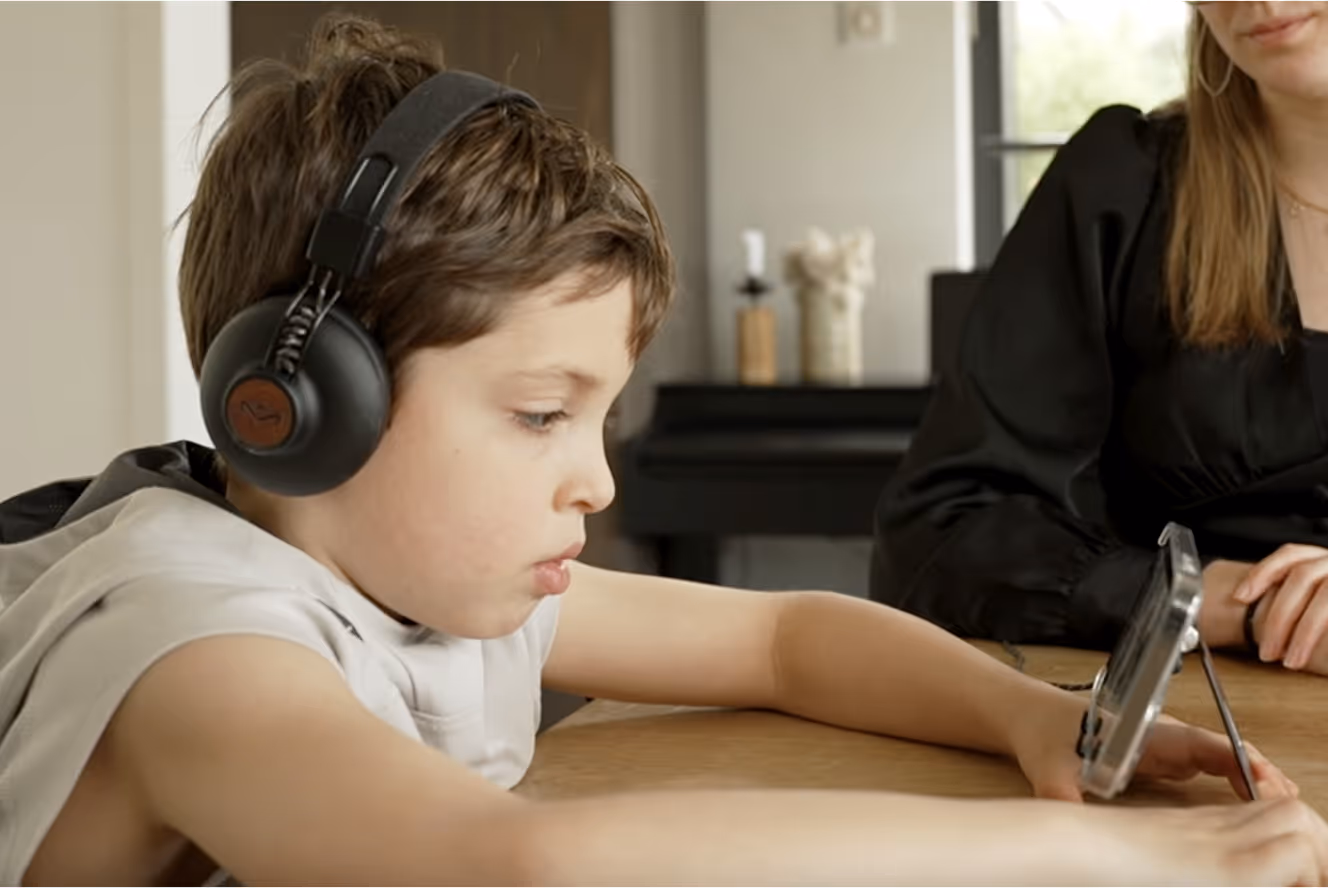 Young boy wearing black headphones looks intently at a smartphone propped up on a table, with a woman sitting nearby.