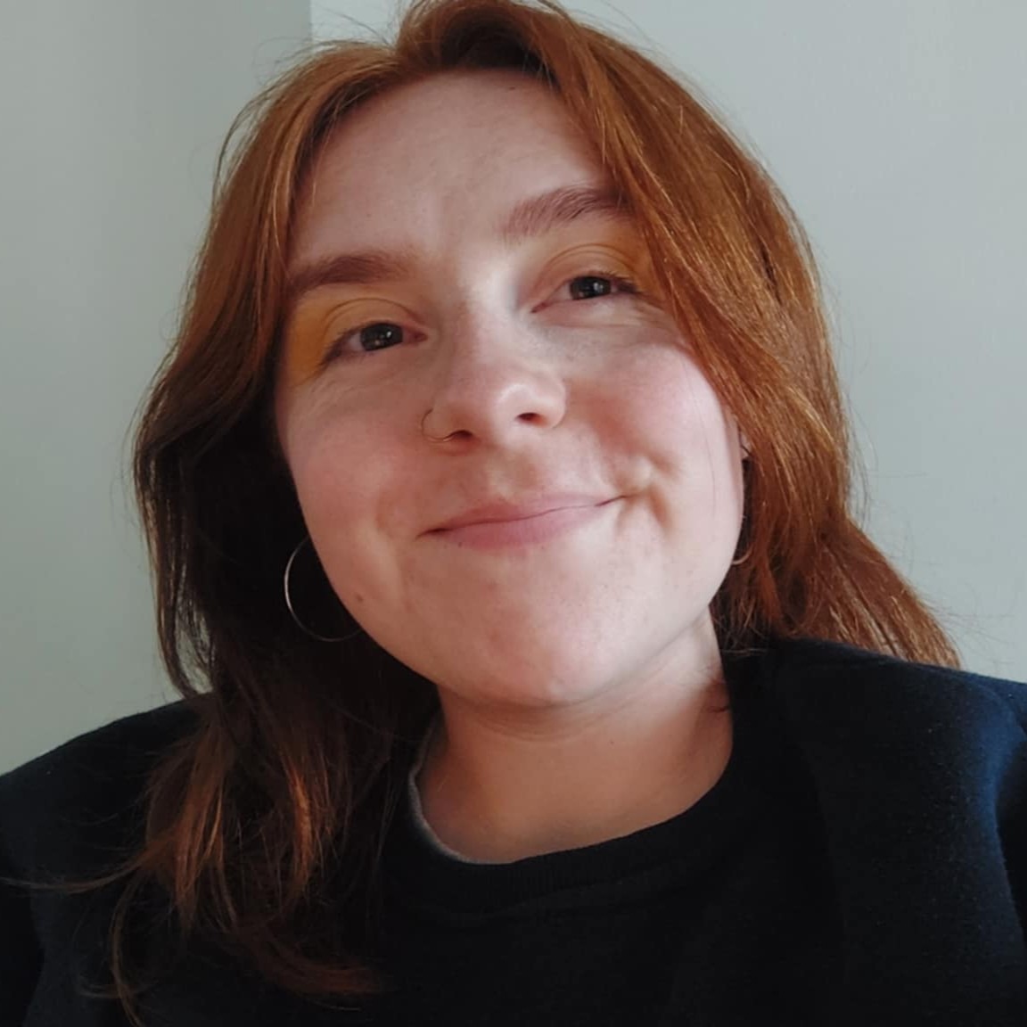 A white person with long wavy ginger hair, smiling, wearing a navy blue sweatshirt, hoop earrings and yellow eyeshadow