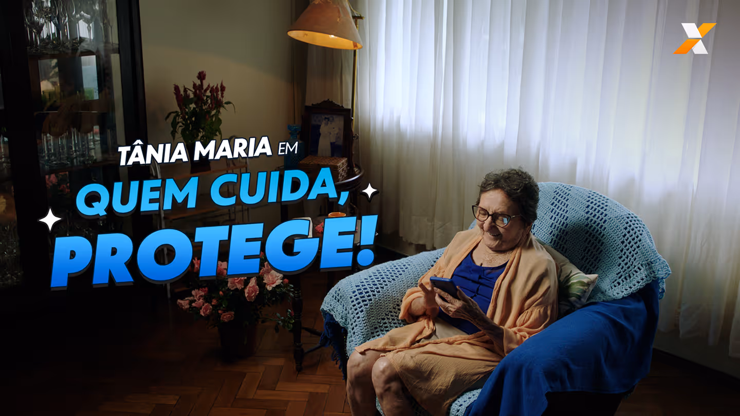 Elderly woman wearing glasses sitting in a blue-covered armchair using a smartphone with flowers and a lamp in the background.