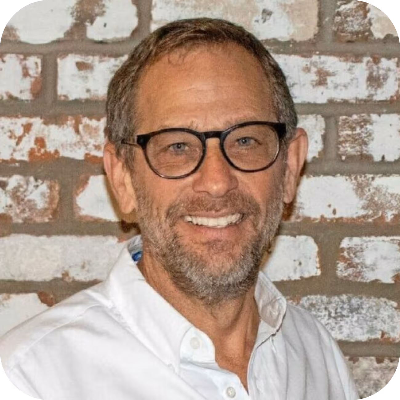 Man smiling while wearing glasses and a white shirt, standing in front of a brick wall background.