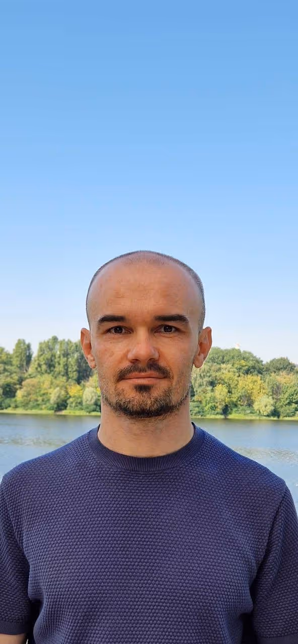 Man standing outdoors wearing a blue sweater, with a lake and lush trees in the background under a clear sky.
