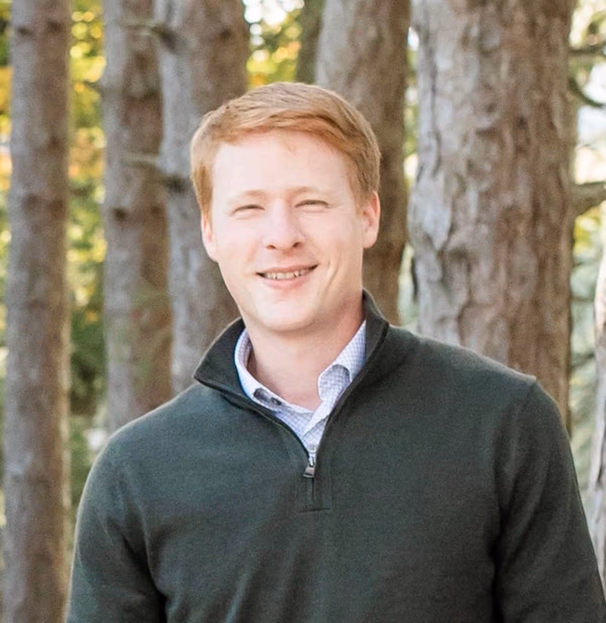 Man smiling while wearing a green sweater and collared shirt, standing outdoors with tall trees in the background.
