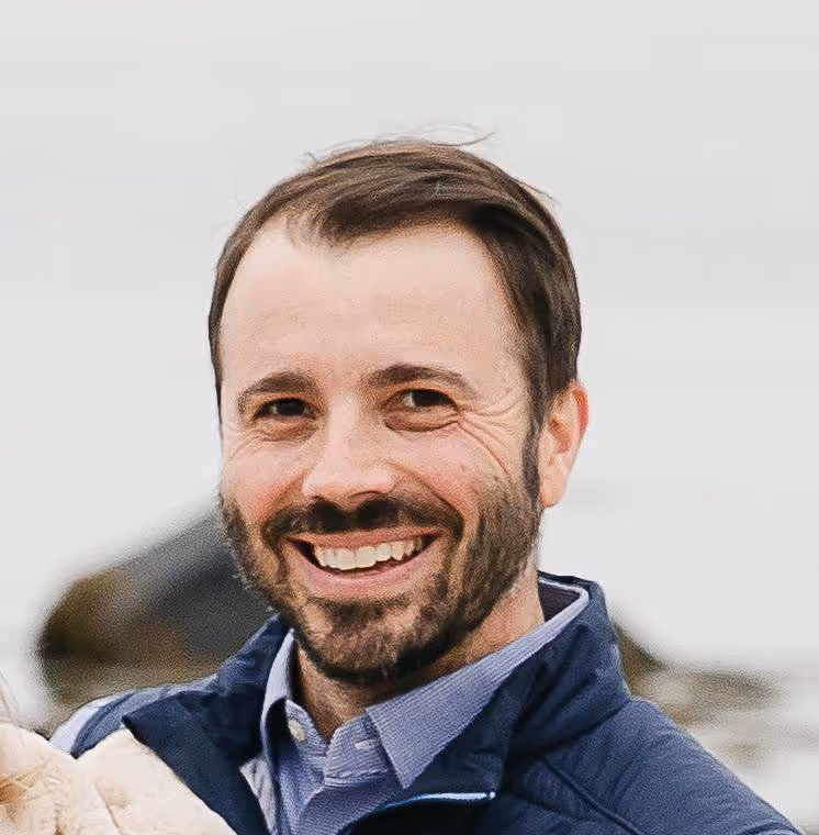 Man smiling while wearing a blue jacket and collared shirt, outdoors with a blurred background of rocks and water.