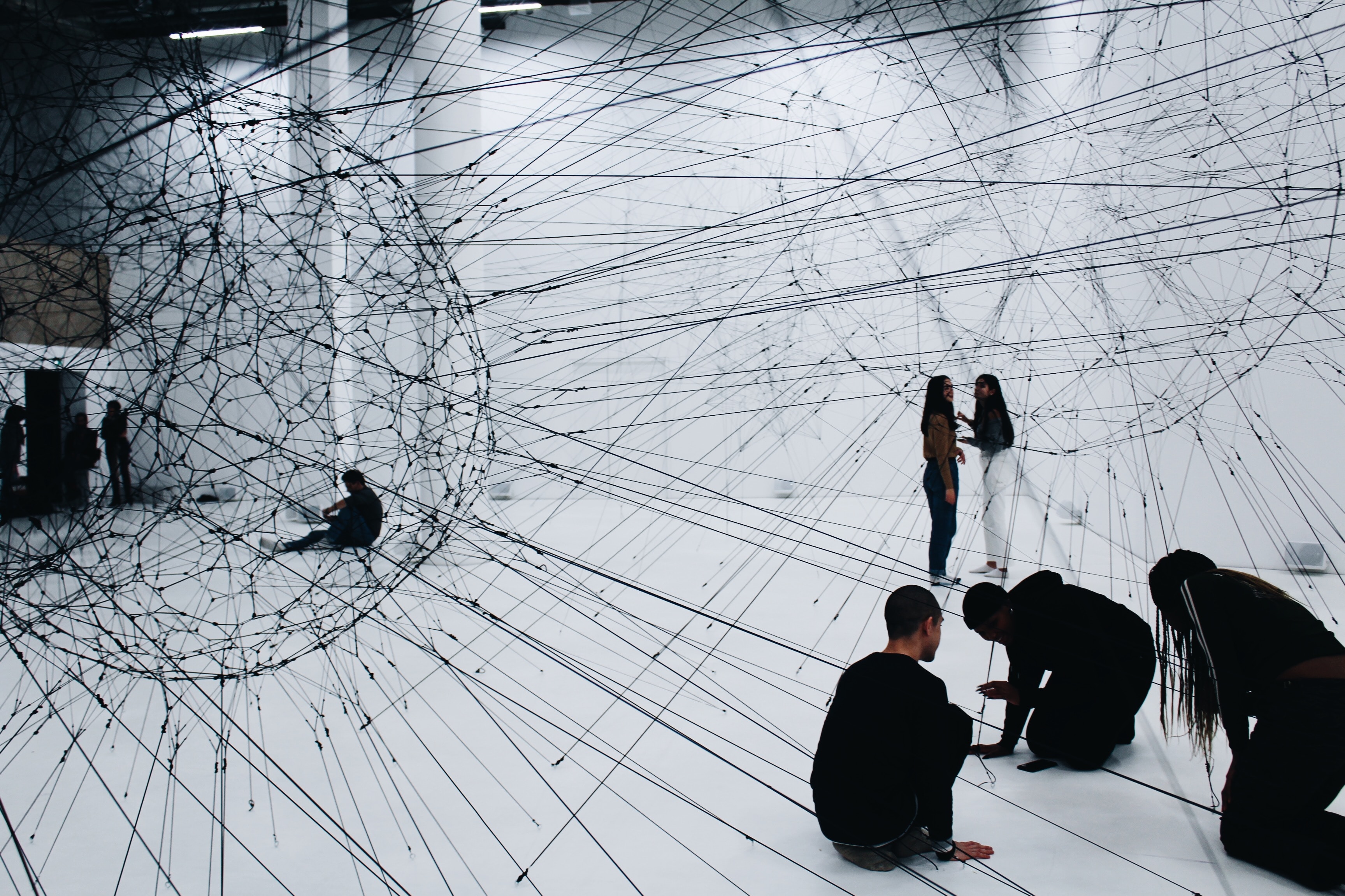People interacting with an art installation made of interconnected black strings forming spheres in a white gallery space.