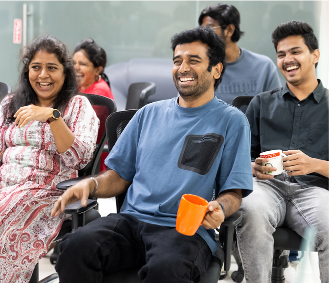 Group of four young adults sitting on office chairs, smiling and laughing, two holding coffee mugs.
