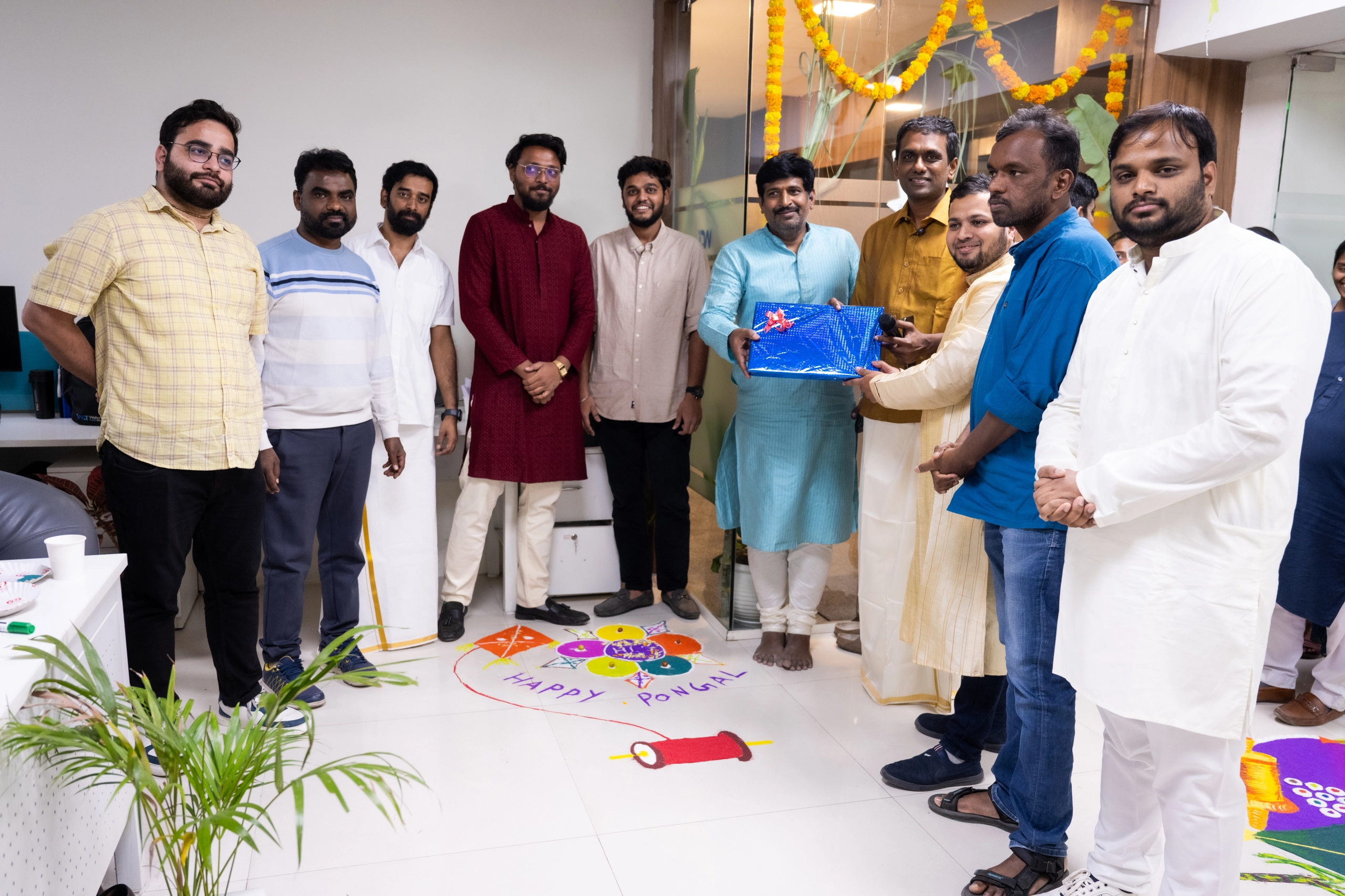Group of men standing indoors around a colorful floor rangoli with text 'Happy Pongal,' one man in a blue kurta holding a wrapped gift box.