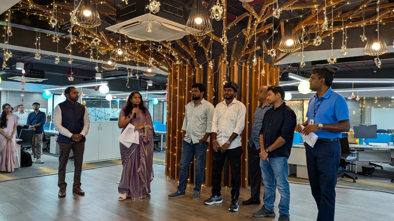 A group of seven people standing in a decorated office space with string lights and wooden pillars, one woman speaking into a microphone.