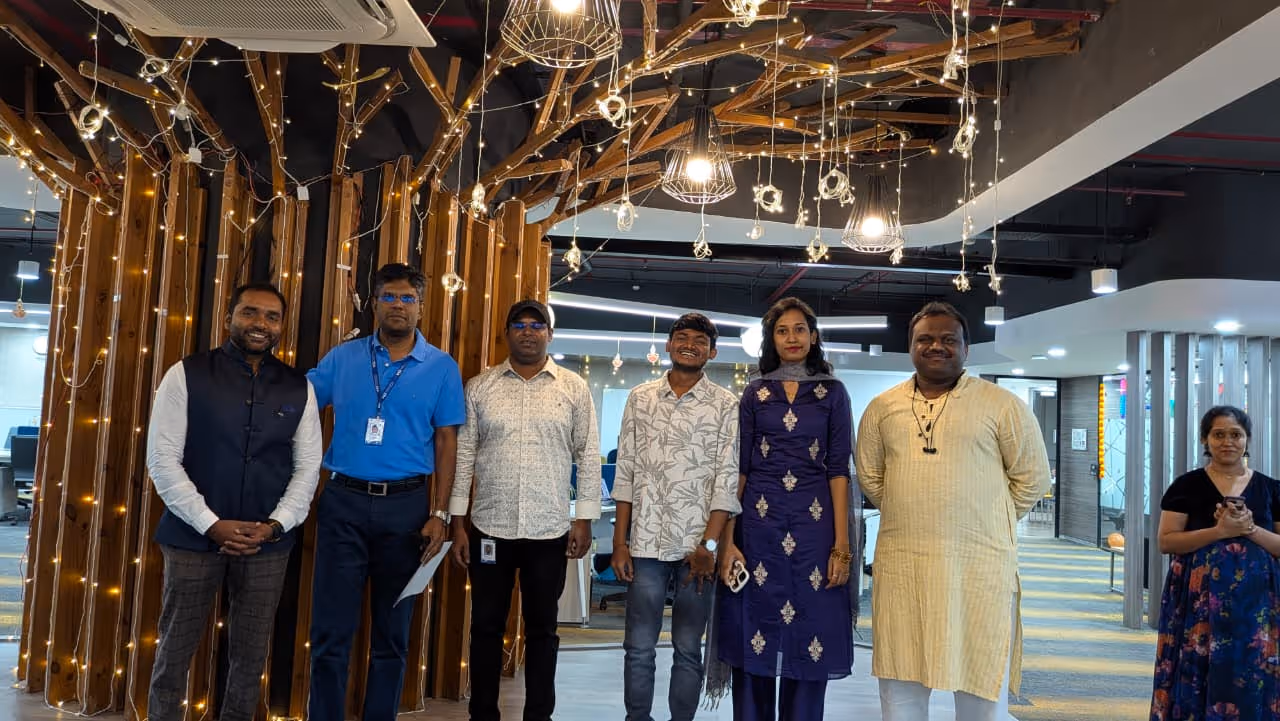 Seven people standing indoors in front of a decorative wooden structure with string lights, smiling and posing for the photo.